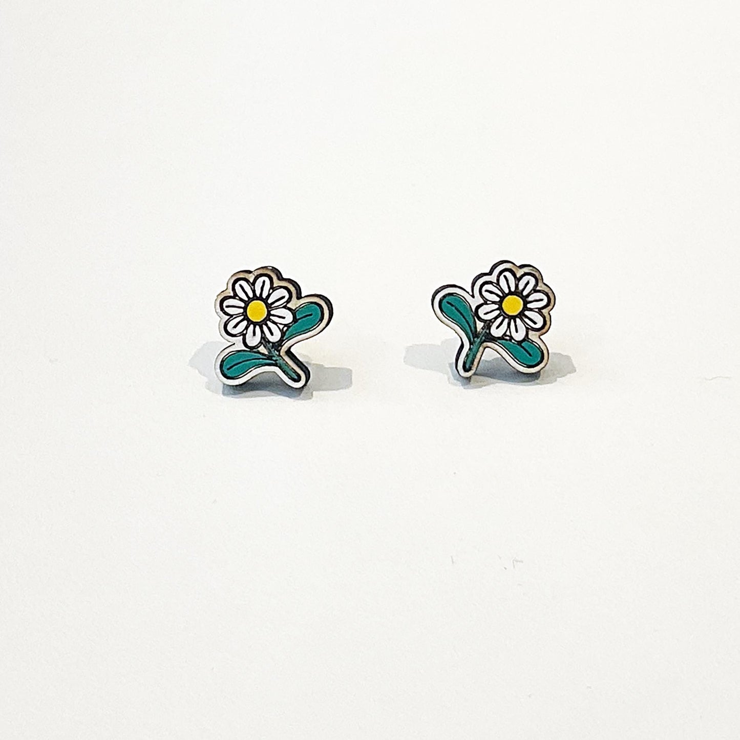 A pair of small, handcrafted stud earrings shaped like daisy flowers. Each earring features white petals with a yellow centre and two green leaves, set against a plain white background. The earrings are symmetrically designed, lightweight, and have a playful, nature-inspired look. Perfect for spring or summer, they add a cute and feminine touch to any outfit, making them ideal for those who love floral or cottagecore-themed accessories.