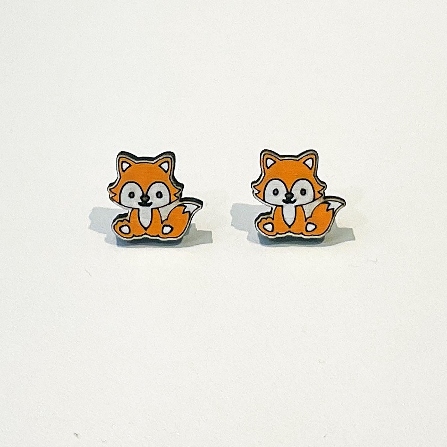 A pair of cute fox-shaped stud earrings with a kawaii design. Each fox has a happy face, orange fur, white belly and cheeks, and a fluffy tail curled beside it. The foxes are sitting upright and facing forward. The earrings have a clean white background to highlight the playful woodland animal design. These earrings are small, lightweight, and perfect for lovers of fun, quirky accessories or forest-themed jewellery.