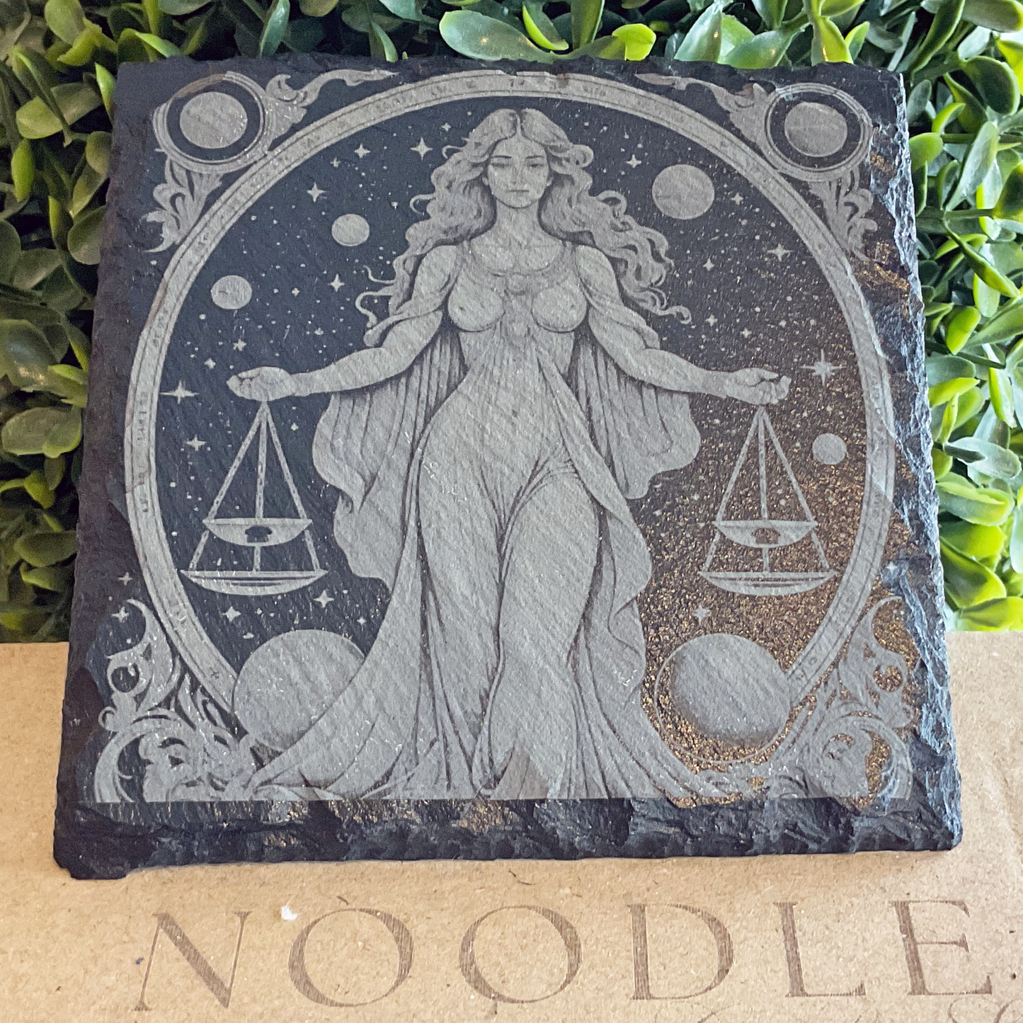 Engraved black slate coaster featuring a detailed illustration of the Libra zodiac symbol—a graceful woman in flowing robes holding a set of balanced scales in each hand. She stands within a circular celestial frame adorned with stars, planets, and swirling ornamental designs. The coaster rests on a cardboard surface with the word "NOODLE" partially visible, surrounded by vibrant green foliage in the background.