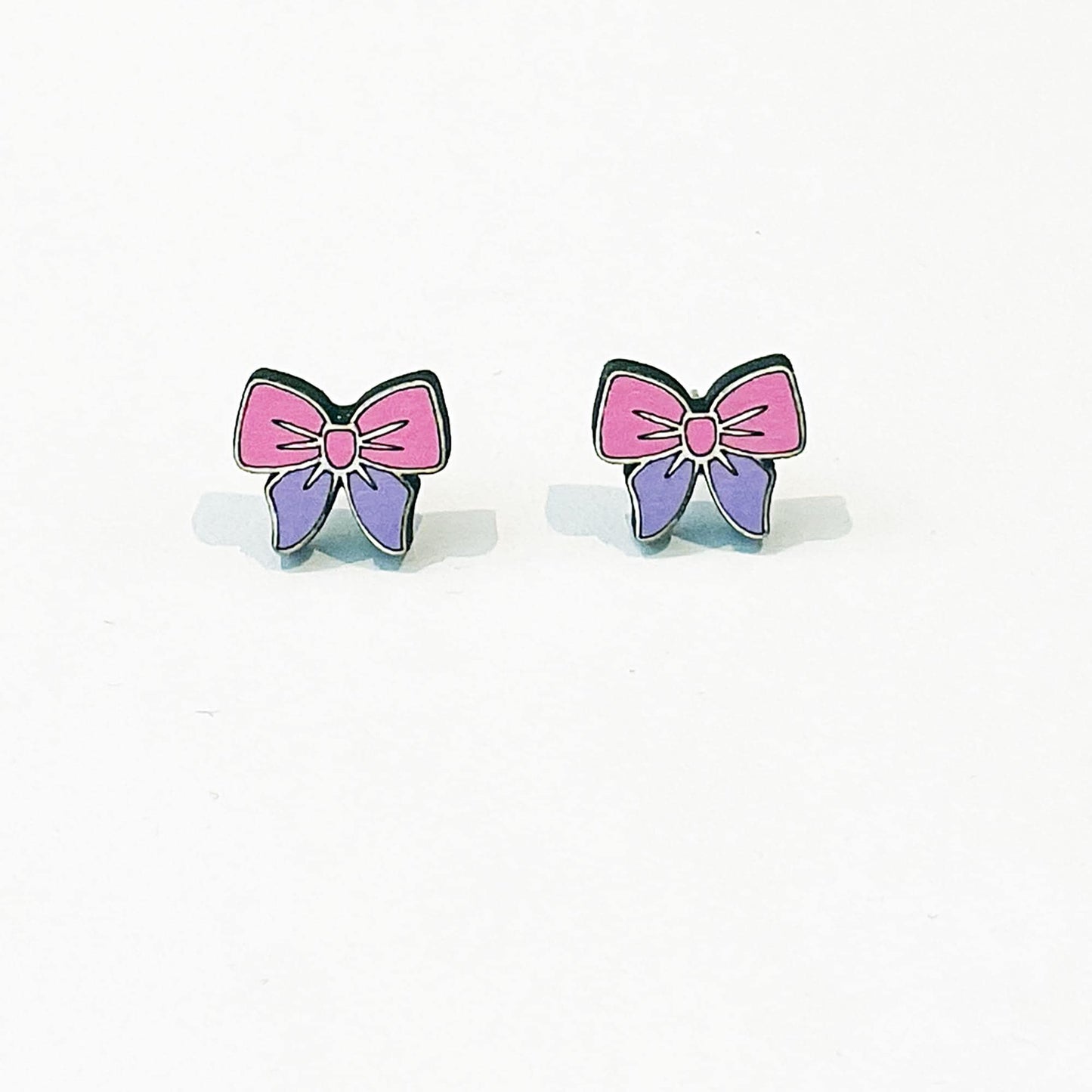 A pair of small, cute stud earrings shaped like bows. Each earring has a pink top half and a purple bottom half, outlined in black, with a pink centre knot. The design is symmetrical and cartoon-like, with a glossy finish. The earrings are displayed upright on a clean white background with a faint shadow beneath each, suggesting a flat surface. These pastel-coloured accessories have a soft, playful aesthetic, perfect for a kawaii or girly fashion style.