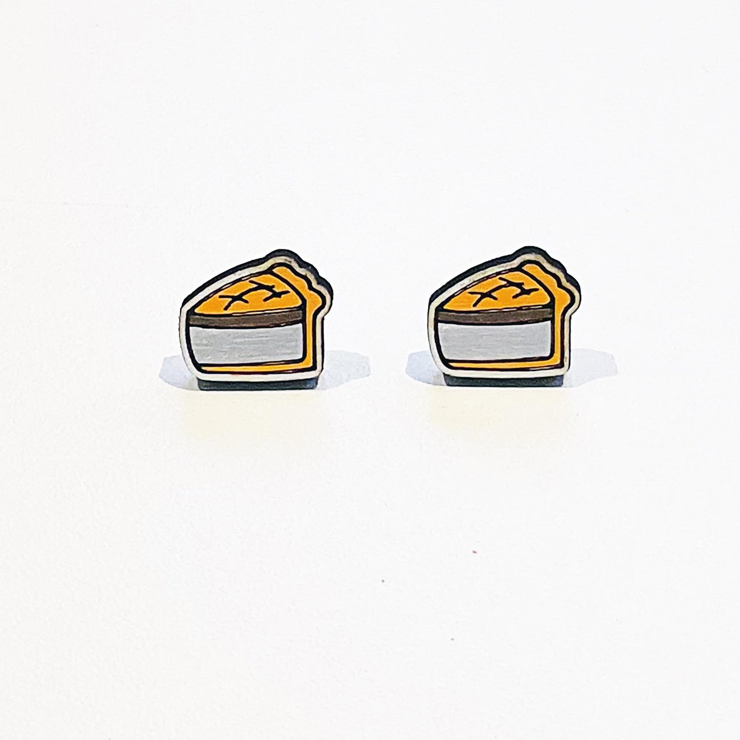 A pair of handmade stud earrings shaped like classic British pork pies. Each earring features a detailed slice of golden-brown pork pie with a crimped pastry edge and visible meat filling, outlined in black for a bold, cartoon-style finish. The design is flat with a glossy surface and stainless steel posts attached at the back. The earrings are displayed against a plain white background with soft shadows beneath them.