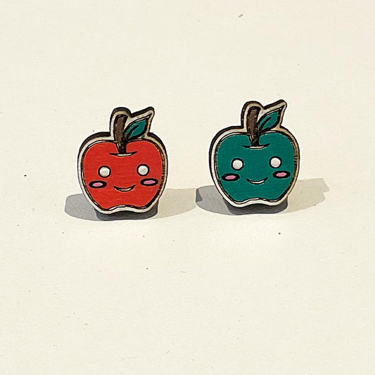 A pair of mismatched apple stud earrings is displayed against a light background. One earring is a red apple, the other is a green apple. Both apples have cute smiling cartoon faces with rosy cheeks and a small green leaf attached to a brown stem. The earrings are made of a lightweight material and have a glossy finish, giving them a playful and cheerful appearance. These quirky, kawaii-style earrings are perfect for fans of fun, food-themed jewellery and handmade accessories.