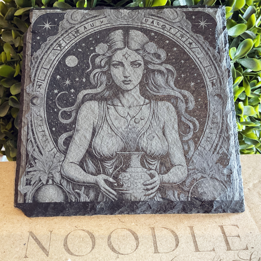 Engraved black slate coaster featuring an intricate illustration of the Aquarius zodiac symbol—a celestial woman with long, wavy hair holding a water vessel. She is surrounded by a circular cosmic frame adorned with stars, moons, and astrological symbols. The border includes detailed botanical and celestial embellishments. The coaster rests on a cardboard surface with the word "NOODLE" partially visible, set against a background of lush green foliage.