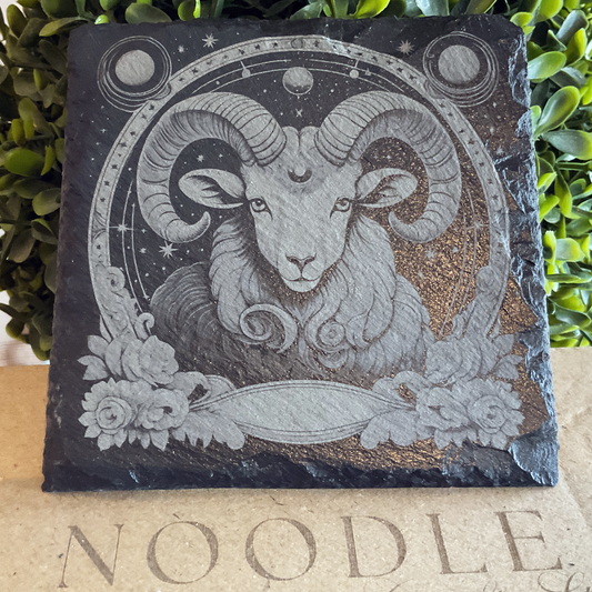 Engraved black slate coaster featuring an intricate illustration of the Aries zodiac symbol—a ram with large curled horns—surrounded by celestial elements like stars, planets, and a crescent moon. Decorative floral accents frame the design, and the coaster rests on a cardboard surface with the word "NOODLE" partially visible. Greenery in the background adds a natural touch.