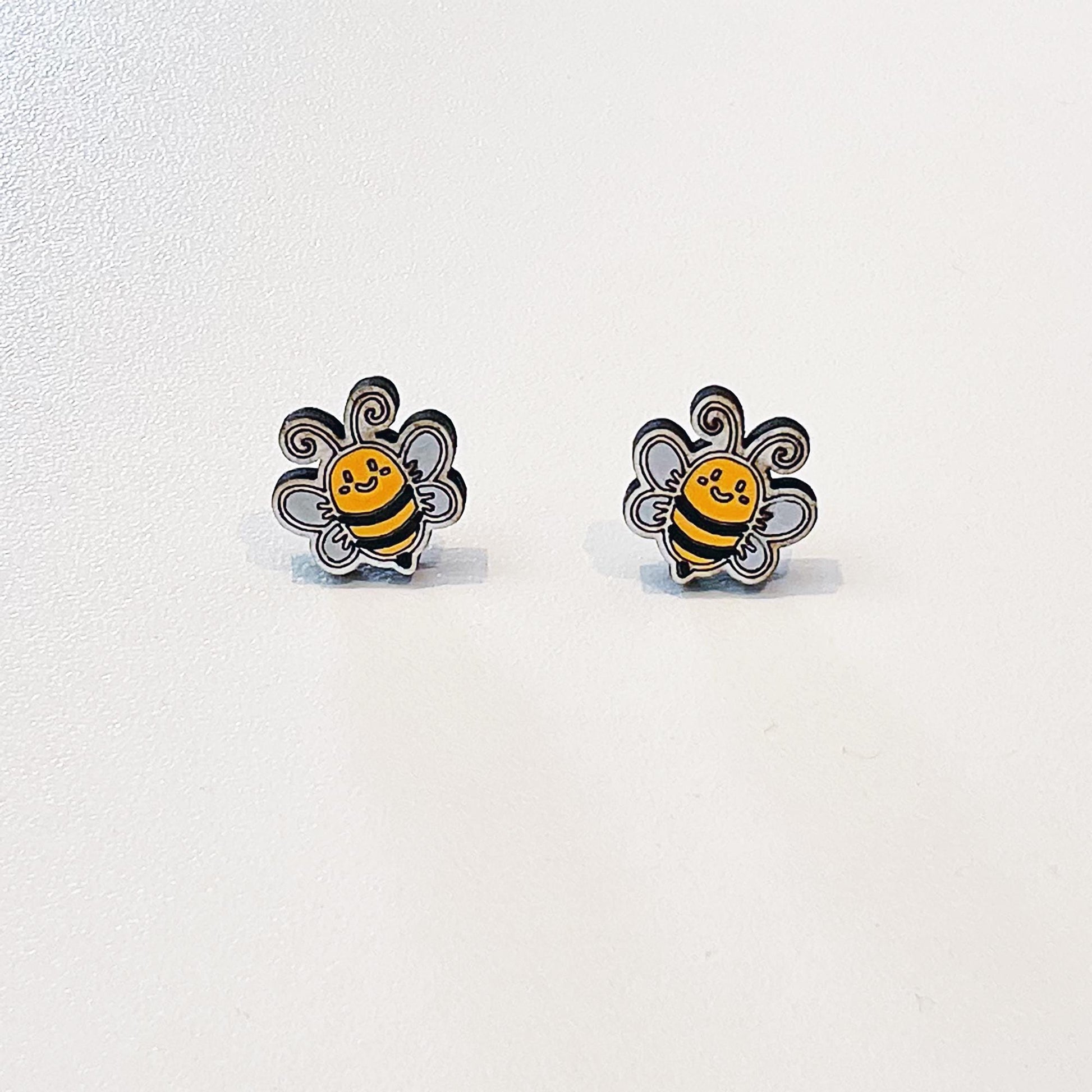 A pair of adorable bumblebee stud earrings shaped like smiling bees with black and yellow stripes, white wings, and curly antennae. Each earring features a happy expression and fine black outlines, giving them a playful, cartoon-like look. Displayed on a white background, these earrings are small, lightweight, and perfect for nature lovers or as a cute, handmade gift.