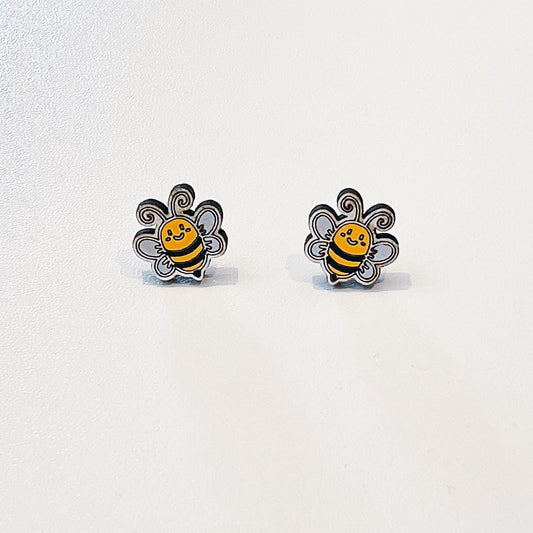 A pair of adorable bumblebee stud earrings shaped like smiling bees with black and yellow stripes, white wings, and curly antennae. Each earring features a happy expression and fine black outlines, giving them a playful, cartoon-like look. Displayed on a white background, these earrings are small, lightweight, and perfect for nature lovers or as a cute, handmade gift.