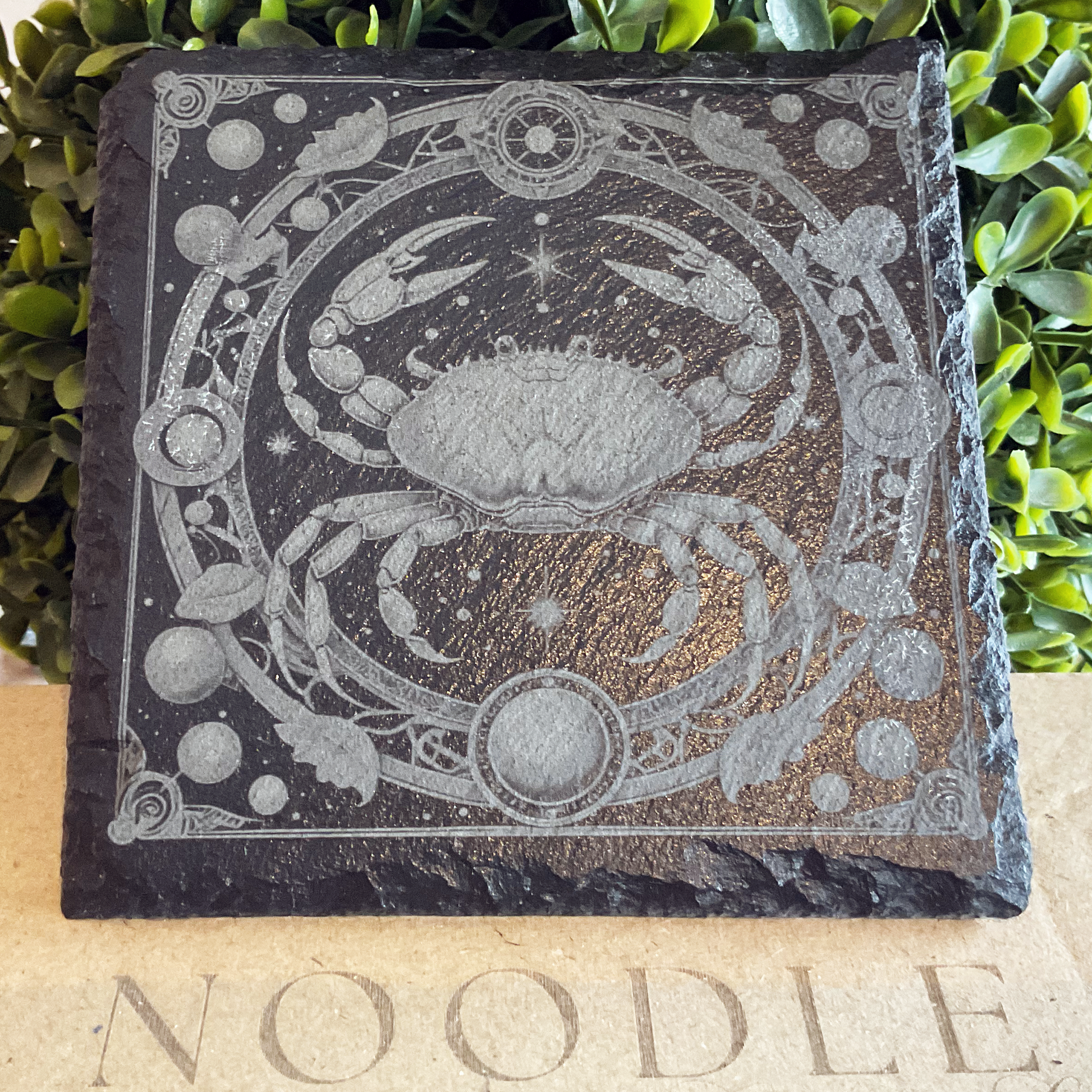 Engraved black slate coaster featuring a detailed illustration of the Cancer zodiac symbol—a stylized crab with raised claws—centered within a circular frame adorned with stars, planets, and botanical elements. The design has a cosmic, mystical aesthetic with intricate linework and ornamental flourishes. The coaster rests on a cardboard surface with the word "NOODLE" partially visible, with lush green foliage in the background.