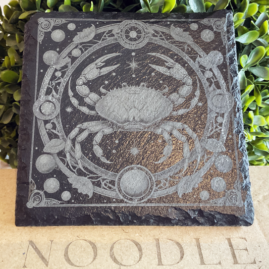 Engraved black slate coaster featuring a detailed illustration of the Cancer zodiac symbol—a stylized crab with raised claws—centered within a circular frame adorned with stars, planets, and botanical elements. The design has a cosmic, mystical aesthetic with intricate linework and ornamental flourishes. The coaster rests on a cardboard surface with the word "NOODLE" partially visible, with lush green foliage in the background.