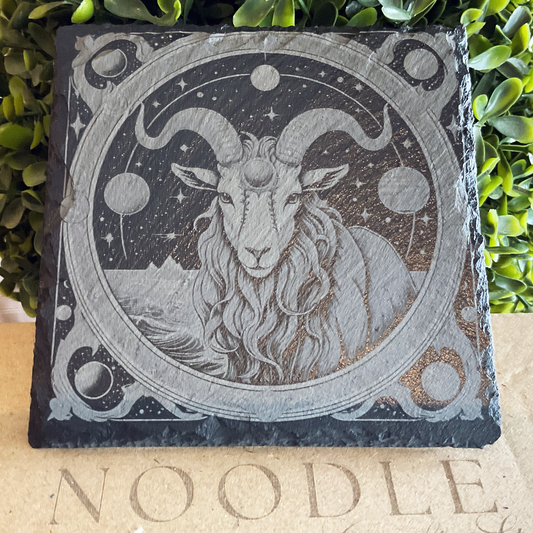 Engraved black slate coaster featuring an intricate illustration of the Capricorn zodiac symbol—a goat with long curled horns and flowing fur—framed by celestial elements including stars, planets, and a crescent moon. The background depicts mountains and a starry night sky. The coaster rests on a cardboard surface with the word "NOODLE" partially visible, with green foliage in the background.