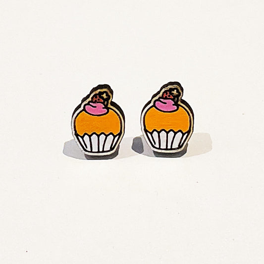 Cupcake Stud EarringsImage shows a pair of handmade stud earrings shaped like cupcakes. Each earring features a golden yellow cupcake base in a white wrapper, topped with pink icing and a small brown cherry with a yellow star accent. The earrings have a cute, cartoon-style design with bold black outlines, giving them a playful and kawaii aesthetic. They are displayed against a white background, making the vibrant colours and charming details stand out clearly.