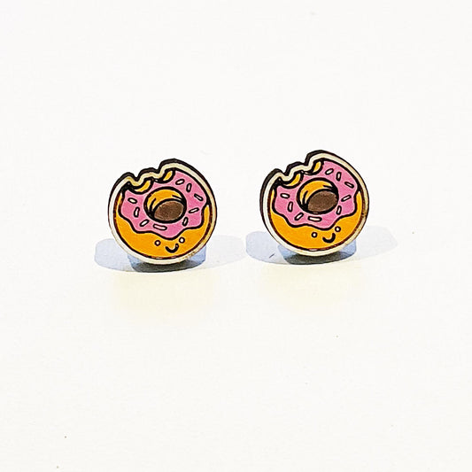 Image shows a pair of novelty stud earrings shaped like pink-frosted doughnuts with a small bite taken out of each. The doughnuts are golden brown with pink icing and sprinkles, featuring a cute smiling face under the hole. They are set against a plain white background, with stainless steel posts for pierced ears. The earrings are playful, brightly coloured, and lightweight – ideal for fun, casual wear or as a quirky gift.