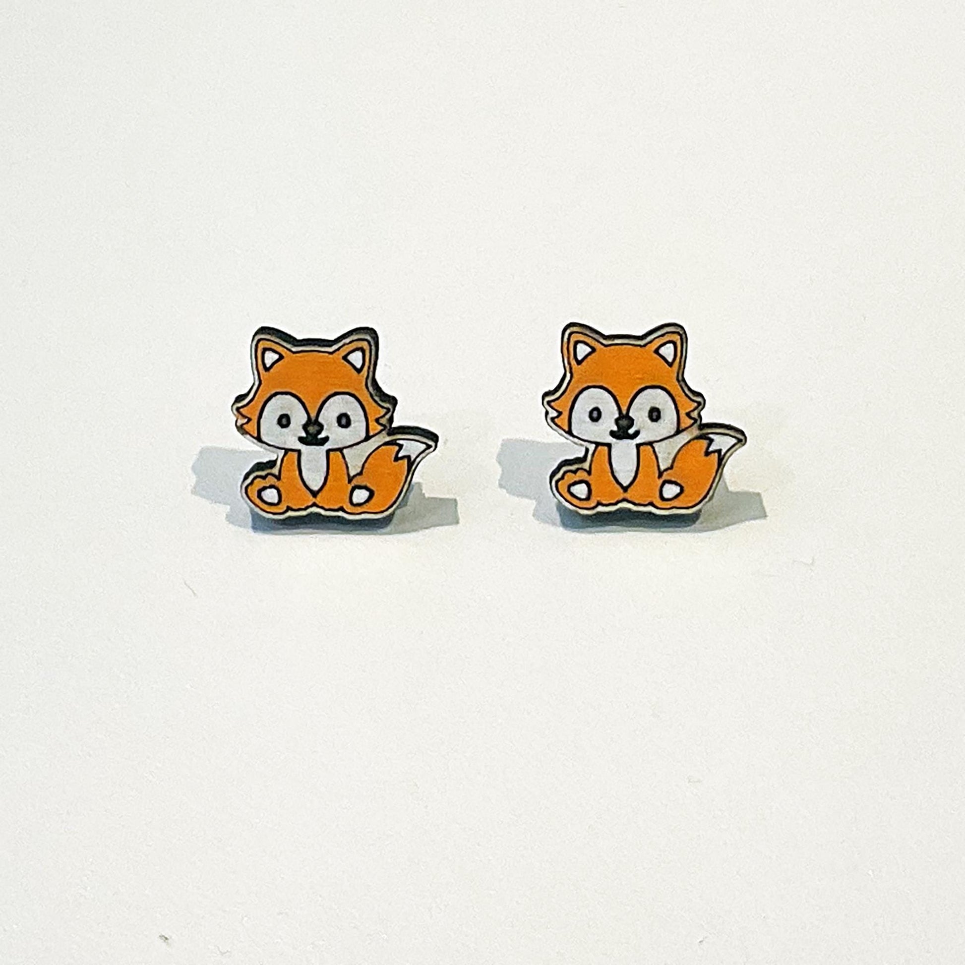 A pair of cute fox-shaped stud earrings with a kawaii design. Each fox has a happy face, orange fur, white belly and cheeks, and a fluffy tail curled beside it. The foxes are sitting upright and facing forward. The earrings have a clean white background to highlight the playful woodland animal design. These earrings are small, lightweight, and perfect for lovers of fun, quirky accessories or forest-themed jewellery.