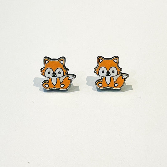 A pair of cute fox-shaped stud earrings with a kawaii design. Each fox has a happy face, orange fur, white belly and cheeks, and a fluffy tail curled beside it. The foxes are sitting upright and facing forward. The earrings have a clean white background to highlight the playful woodland animal design. These earrings are small, lightweight, and perfect for lovers of fun, quirky accessories or forest-themed jewellery.