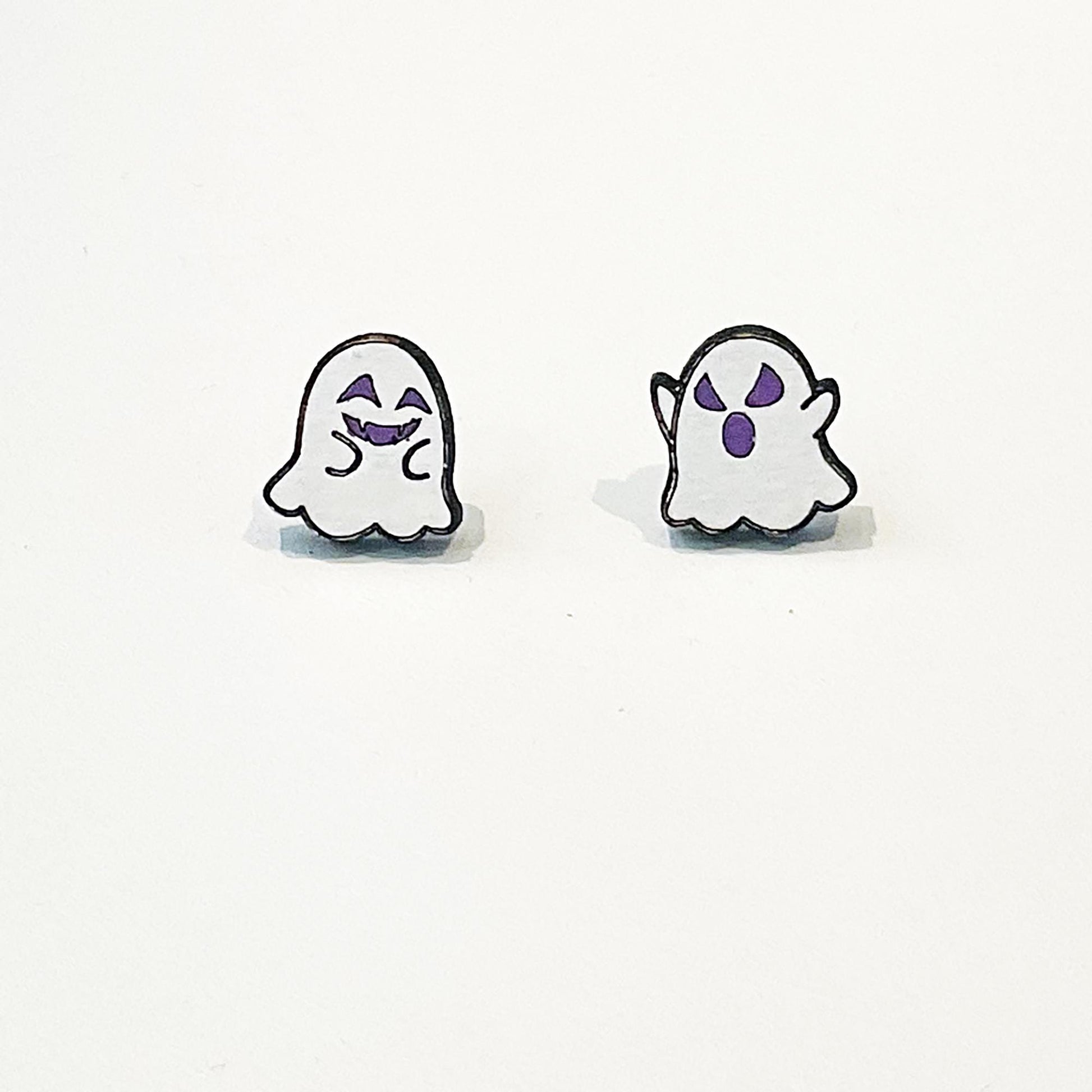 A pair of mismatched ghost-themed stud earrings featuring playful cartoon-style designs. One earring shows a smiling white ghost with closed, happy eyes and a cheeky grin in purple. The other displays a surprised ghost with raised arms and wide, startled purple eyes and mouth. Both ghosts have a simple rounded shape with scalloped bottoms and are outlined in black. The earrings are lightweight, flat, and appear to be made of a glossy material, ideal for Halloween wear.