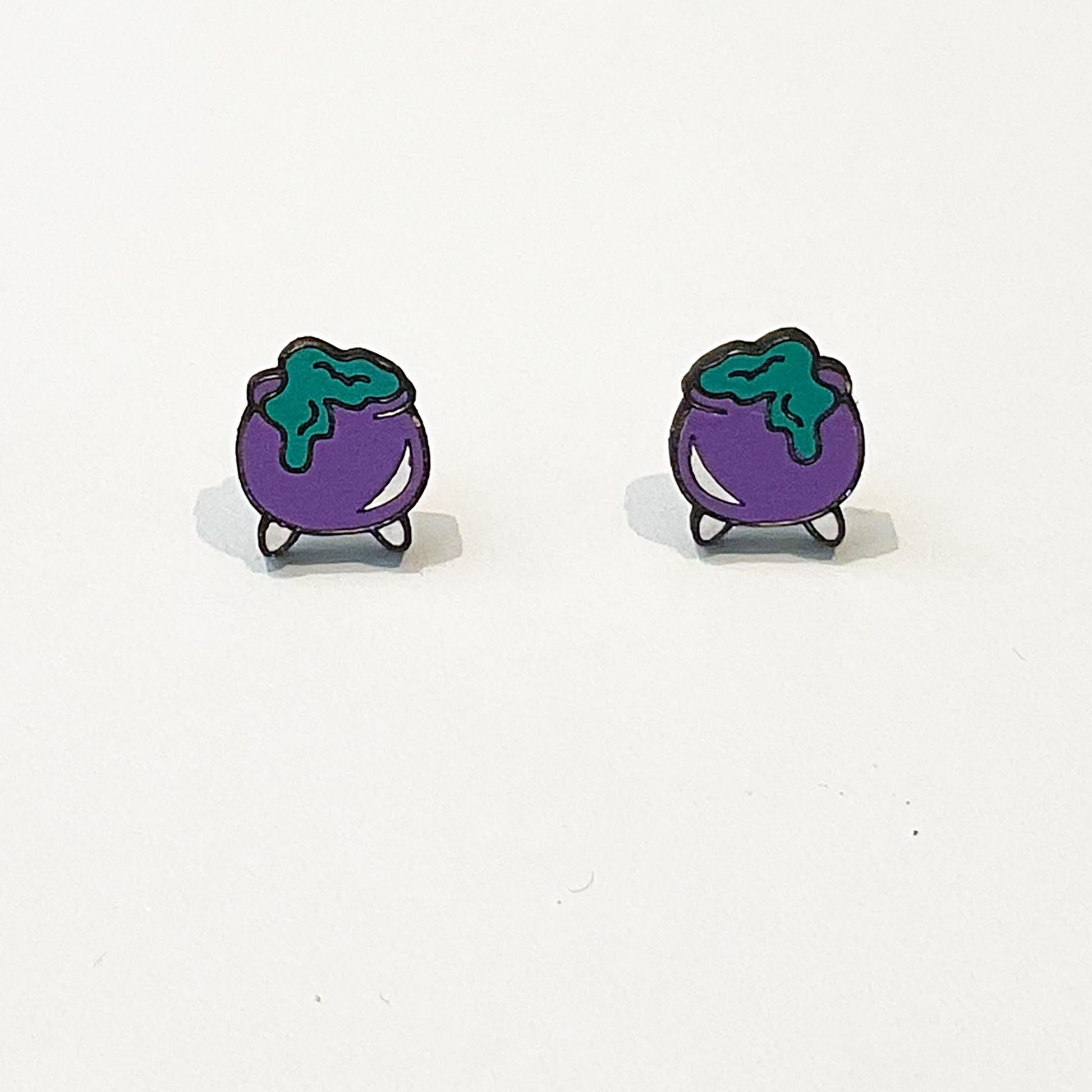 A pair of handmade stud earrings shaped like purple witch’s cauldrons with green bubbling potion overflowing from the top. The cauldrons have a rounded body with small black legs and are outlined in black for definition. The green potion appears to drip slightly down the sides, creating a fun, spooky effect. The earrings are photographed upright on a white background, showing their playful Halloween-inspired design and cartoon-style charm.