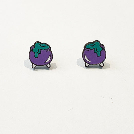 A pair of handmade stud earrings shaped like purple witch’s cauldrons with green bubbling potion overflowing from the top. The cauldrons have a rounded body with small black legs and are outlined in black for definition. The green potion appears to drip slightly down the sides, creating a fun, spooky effect. The earrings are photographed upright on a white background, showing their playful Halloween-inspired design and cartoon-style charm.