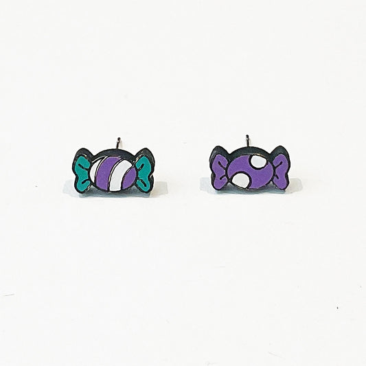 A pair of quirky stud earrings shaped like wrapped sweets, each featuring a unique design. One earring shows a purple and white striped sweet with teal twist ends, while the other displays a purple sweet with white polka dots and purple ends. Both have a playful, cartoon-style look with bold black outlines. The earrings are set against a plain white background and have visible earring posts, highlighting their stud style.