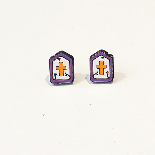 A pair of Halloween-themed stud earrings shaped like cartoon tombstones. Each earring features a white gravestone with a bold orange cross in the centre and small black vine details on either side. The tombstones are outlined in dark purple with a slightly uneven, spooky top edge to give a worn, gothic look. The earrings are flat, lightweight, and have a matte finish, designed in a fun and quirky style perfect for Halloween or gothic fashion lovers. Set against a white background.