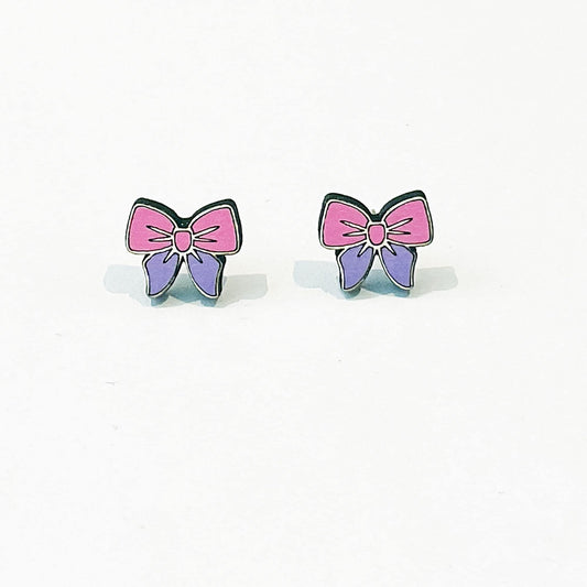 A pair of small, cute stud earrings shaped like bows. Each earring has a pink top half and a purple bottom half, outlined in black, with a pink centre knot. The design is symmetrical and cartoon-like, with a glossy finish. The earrings are displayed upright on a clean white background with a faint shadow beneath each, suggesting a flat surface. These pastel-coloured accessories have a soft, playful aesthetic, perfect for a kawaii or girly fashion style.