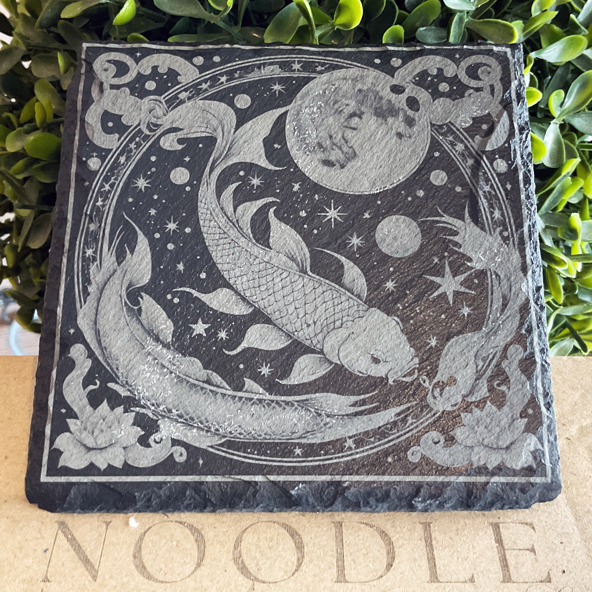 Engraved black slate coaster featuring a detailed illustration of the Pisces zodiac symbol—two koi fish swimming in a circular motion amidst a celestial background filled with stars, moons, and planets. The design includes swirling water elements and blooming lotus flowers, all enclosed in an ornate circular border. The coaster rests on a cardboard surface with the word "NOODLE" partially visible, with green foliage in the background.