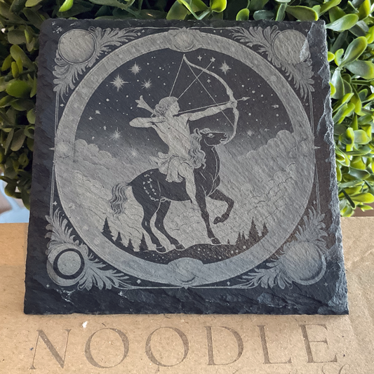 Engraved black slate coaster featuring a detailed illustration of the Sagittarius zodiac symbol—a centaur archer aiming a drawn bow and arrow toward the starry sky. The scene is framed within an ornate circular border, with celestial elements like stars, moons, and clouds, as well as a landscape of pine trees in the background. The coaster rests on a cardboard surface with the word "NOODLE" partially visible, and green foliage in the background.