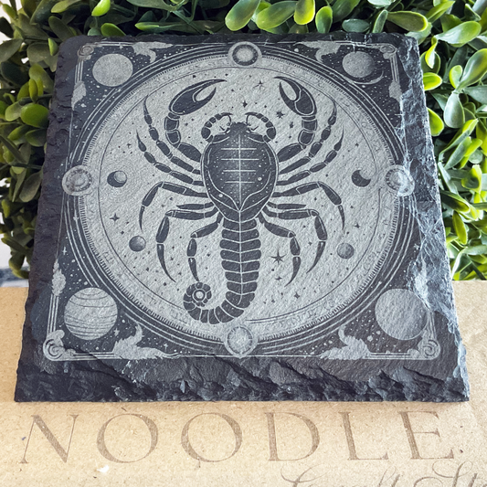 Engraved black slate coaster featuring a striking illustration of the Scorpio zodiac symbol—a detailed scorpion centered within a celestial circular frame. The design includes stars, planets, moons, and ornate cosmic patterns etched around the border. The coaster rests on a cardboard surface with the word "NOODLE" partially visible, with lush green foliage in the background.