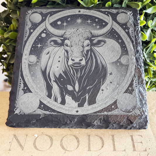 Engraved black slate coaster featuring a powerful illustration of the Taurus zodiac symbol—a strong bull with large, curved horns—standing beneath a star-filled sky. The bull is centered within an ornate circular frame adorned with planets, moons, and celestial motifs. The coaster rests on a cardboard surface with the word "NOODLE" partially visible, with vibrant green foliage in the background.