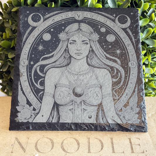 Engraved black slate coaster featuring a detailed illustration of the Virgo zodiac symbol—a serene woman with long flowing hair adorned with floral accents, wearing a celestial-themed outfit with a crescent moon at her chest. She is encircled by a cosmic frame decorated with stars, planets, astrological symbols, and intricate linework. The coaster rests on a cardboard surface with the word "NOODLE" partially visible, against a backdrop of green foliage.